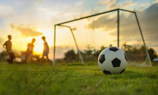 Fußball im Vordergrund, im Hintergrund verschwommen die Umrisse von einem Tor und spielenden Kindern